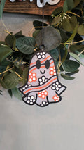 Handmade ghost-shaped car freshie with orange and black floral accents and a smiling face hanging from a rearview mirror.